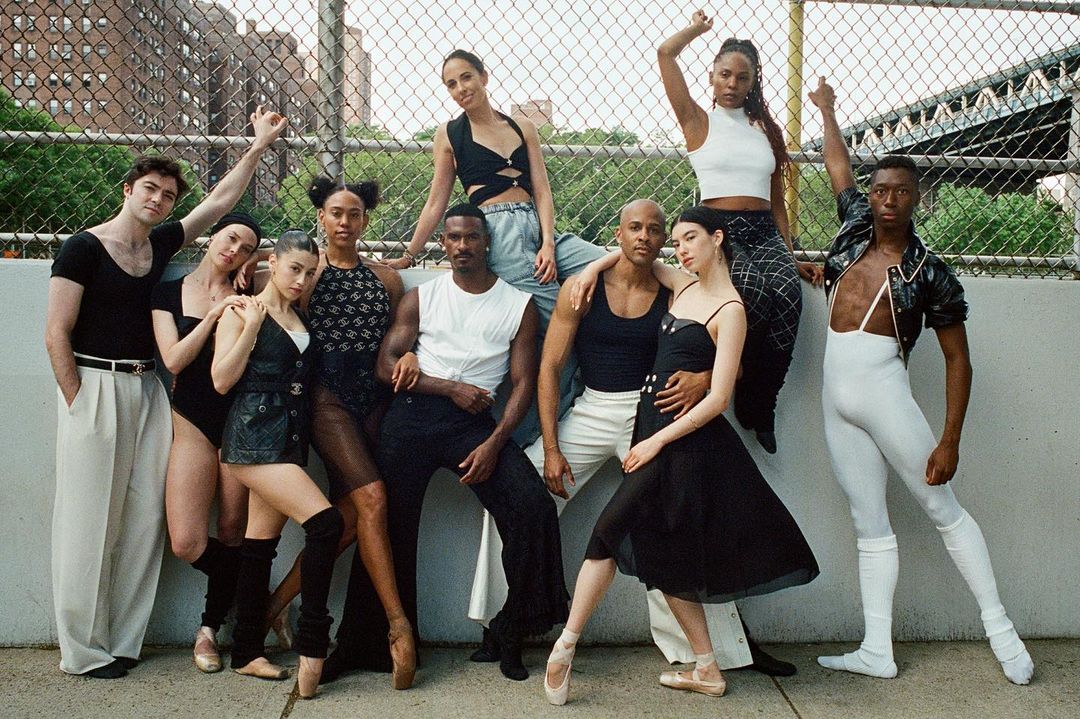 Group of nine ballet dancers posing in black and white athletic wear, outdoors on a stage in front of a fence. 