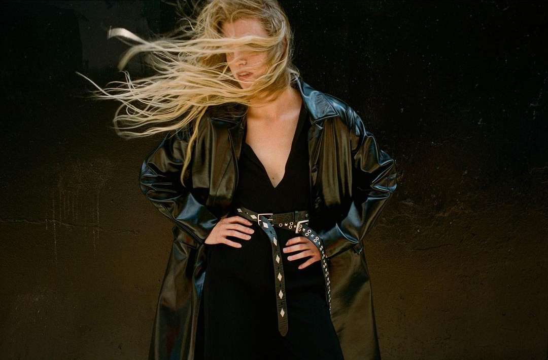 Blonde model in a black dress and trench coat poses against a dark wall, her hair blowing across her face in a fashion-forward image.
