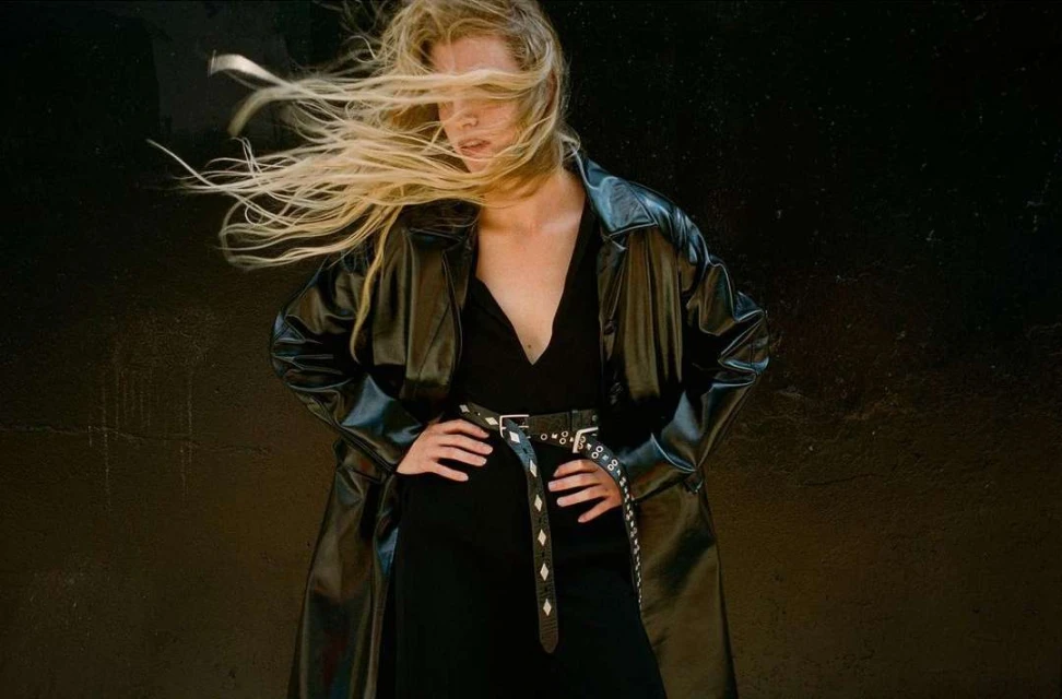 Blonde model in a black dress and trench coat poses against a dark wall, her hair blowing across her face in a fashion-forward image.