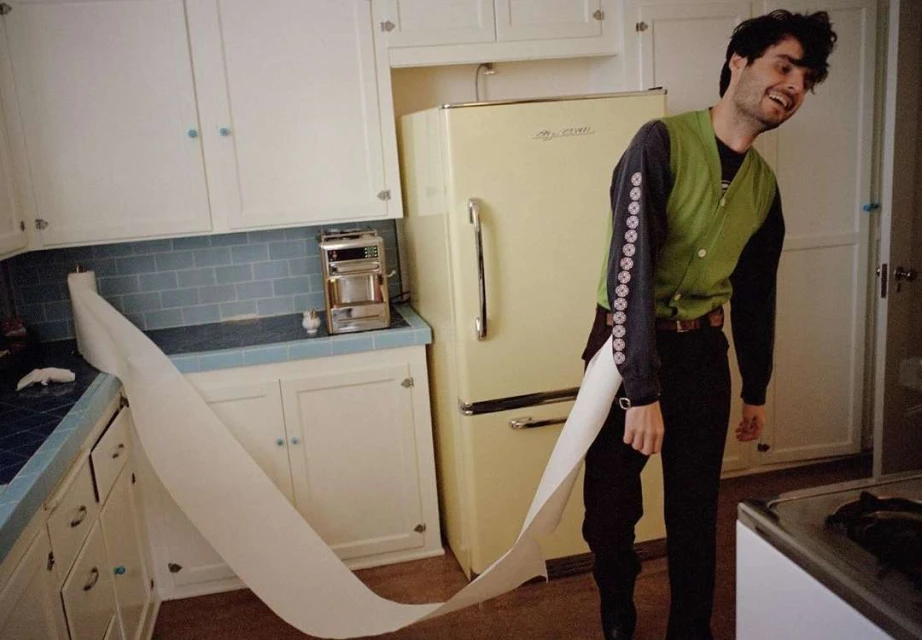 Man in a green vest stands in a kitchen with a long piece of paper trailing from the counter, smiling in a candid, indoor shot.