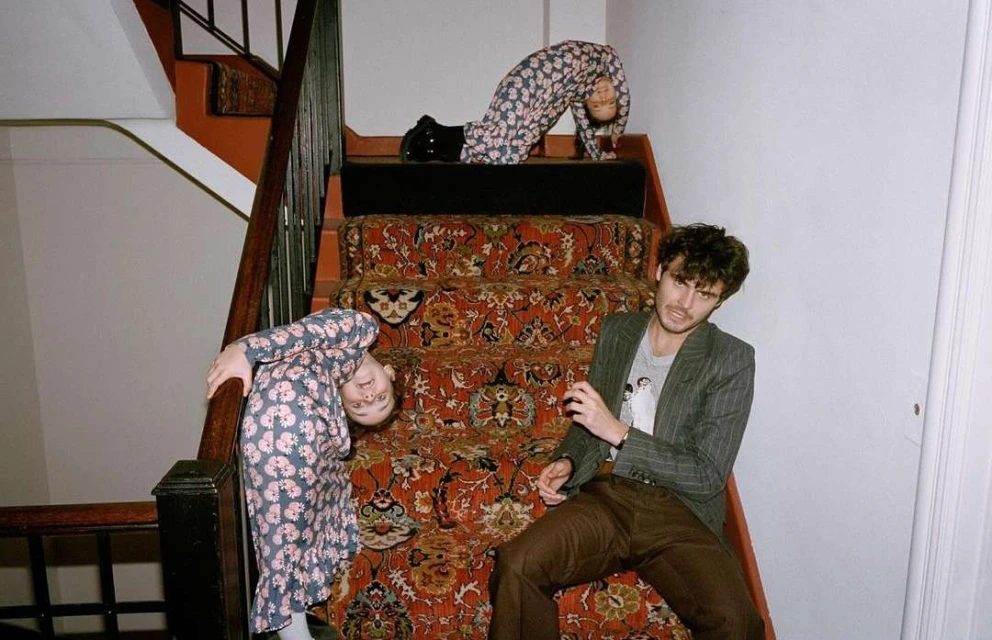 Three individuals creatively pose on a carpeted staircase indoors. One man sits, while two others, a child and an adult, are upside down.