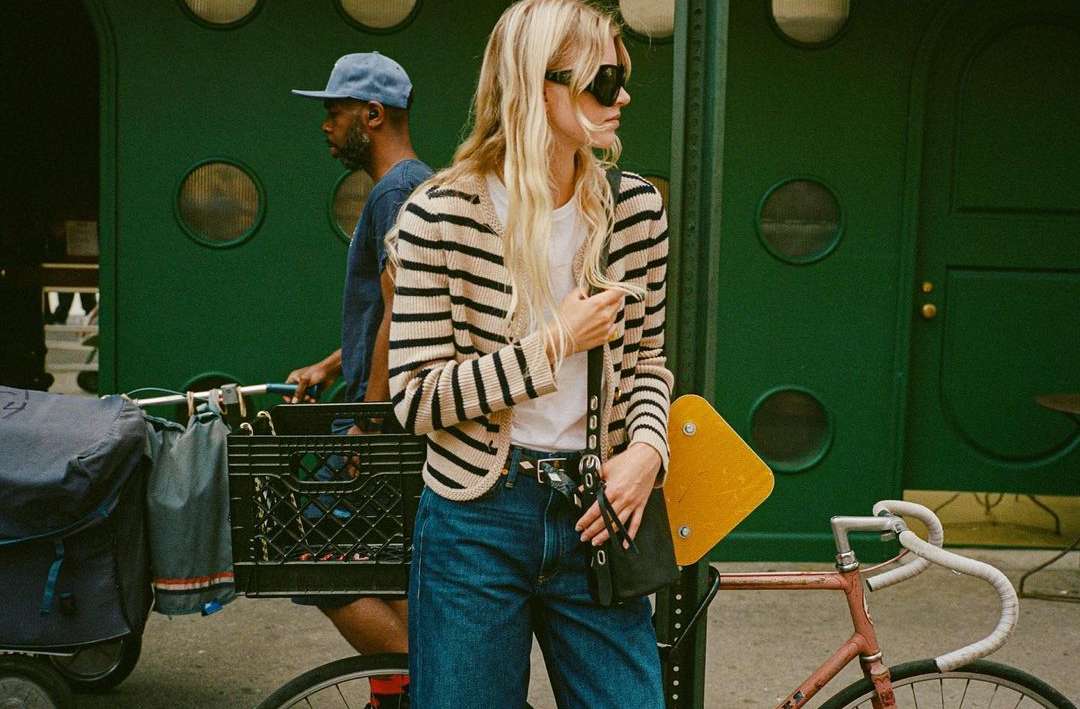 A stylish woman in a striped cardigan, jeans, and sunglasses stands next to a bicycle on an urban street with a man in the background.