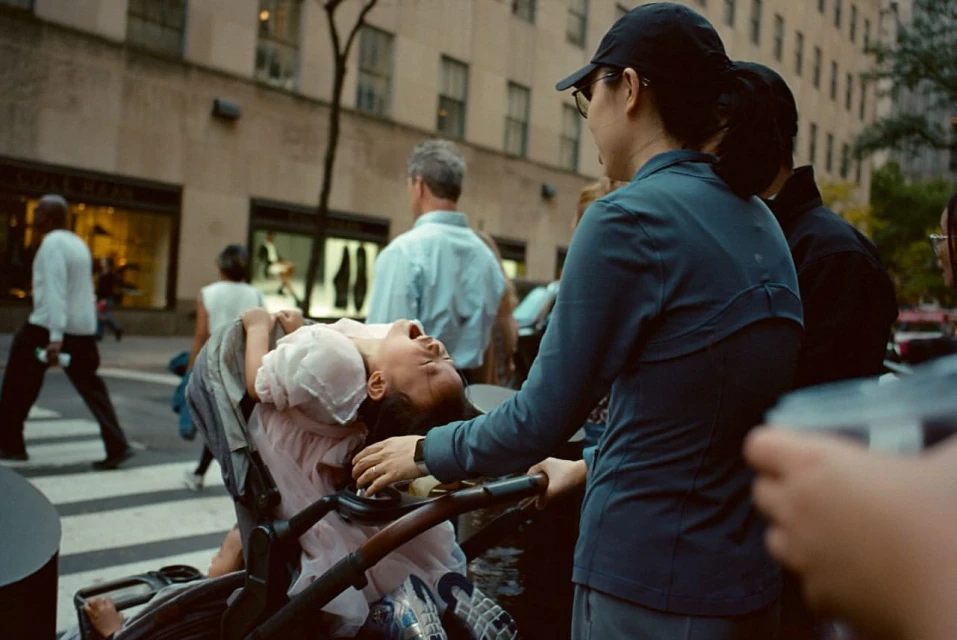 A toddler in a stroller is being attended to by an adult on a city street with pedestrians and storefronts in the background.