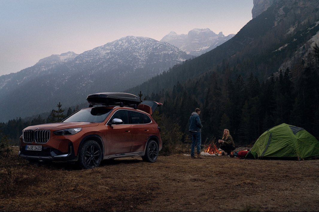 Orange BMW SUV with cargo carrier at a campsite in front of a mountain range with a couple sitting by a campfire near a tent.