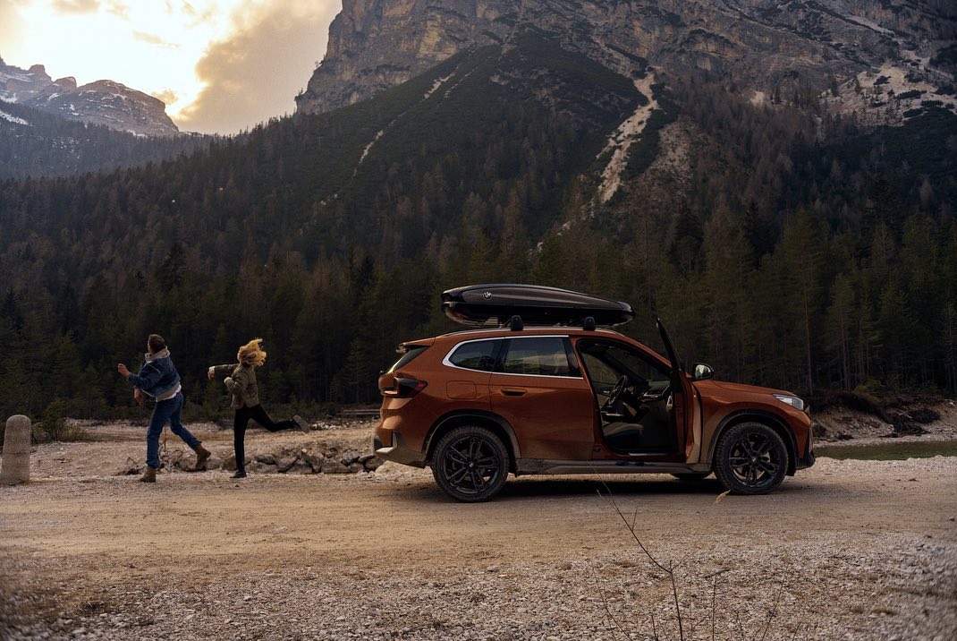 Orange SUV parked with open door in mountainous landscape with two people, one running, and a backdrop of trees and mountain peaks.