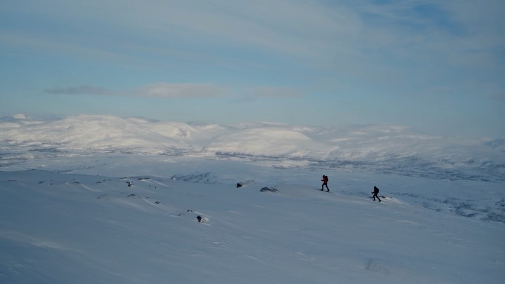Two people hike through a snow-covered mountain landscape under a partly cloudy sky, creating a sense of adventure and exploration.