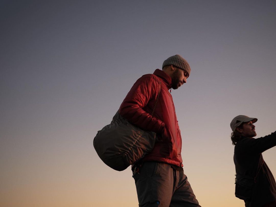 Two adult males stand outdoors; one wears a beanie and red jacket, carrying a bag; the other wears a cap; the sky is clear.