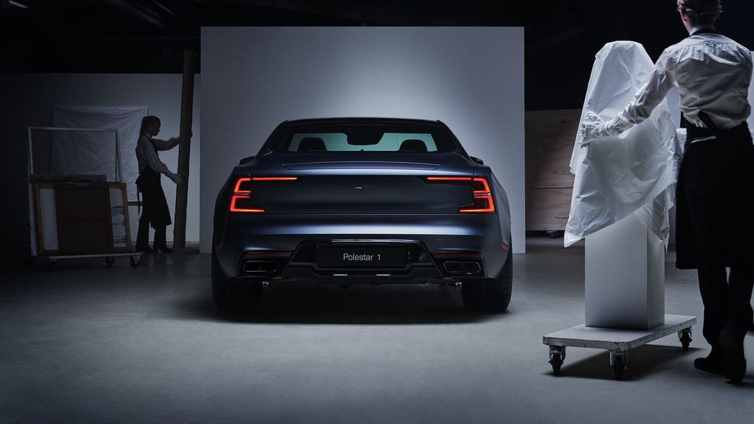 Rear view of a dark blue Polestar 1 car in a photo studio, showcasing its sleek design and taillights, with two people in the background.