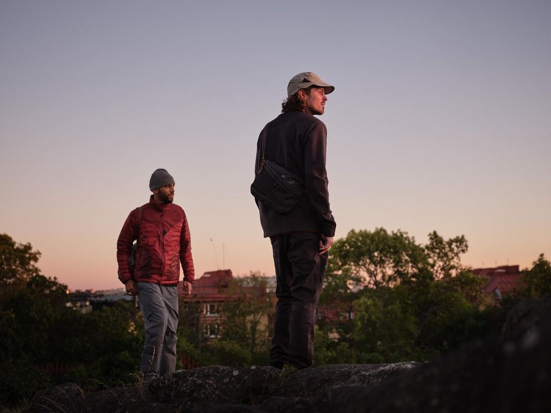 Two men stand on a rocky hilltop with trees and rooftops visible at sunset, wearing casual outdoor attire and looking towards the horizon.