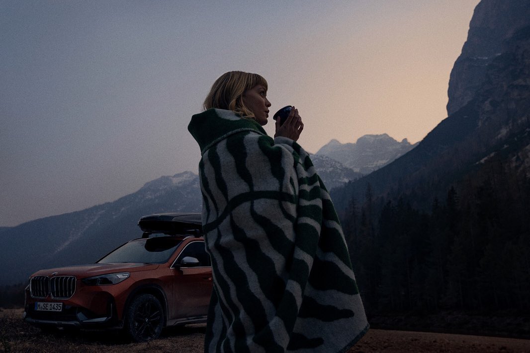 Woman in blanket holding cup stands near orange BMW SUV against mountain backdrop at dusk. Automotive lifestyle, nature, outdoors.