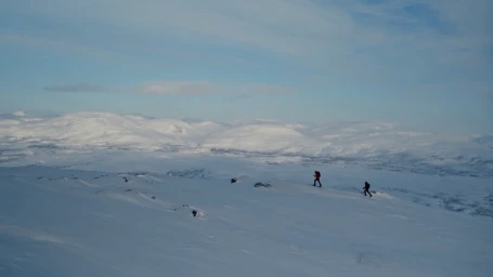 Two hikers walking across a vast snowfield beneath a pale blue sky in a wide landscape photographed by Daniel Blom.