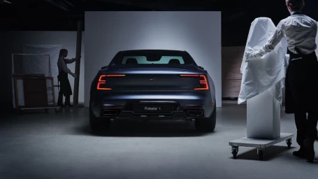 Rear view of a dark coupe car parked in a studio with two people working around it.