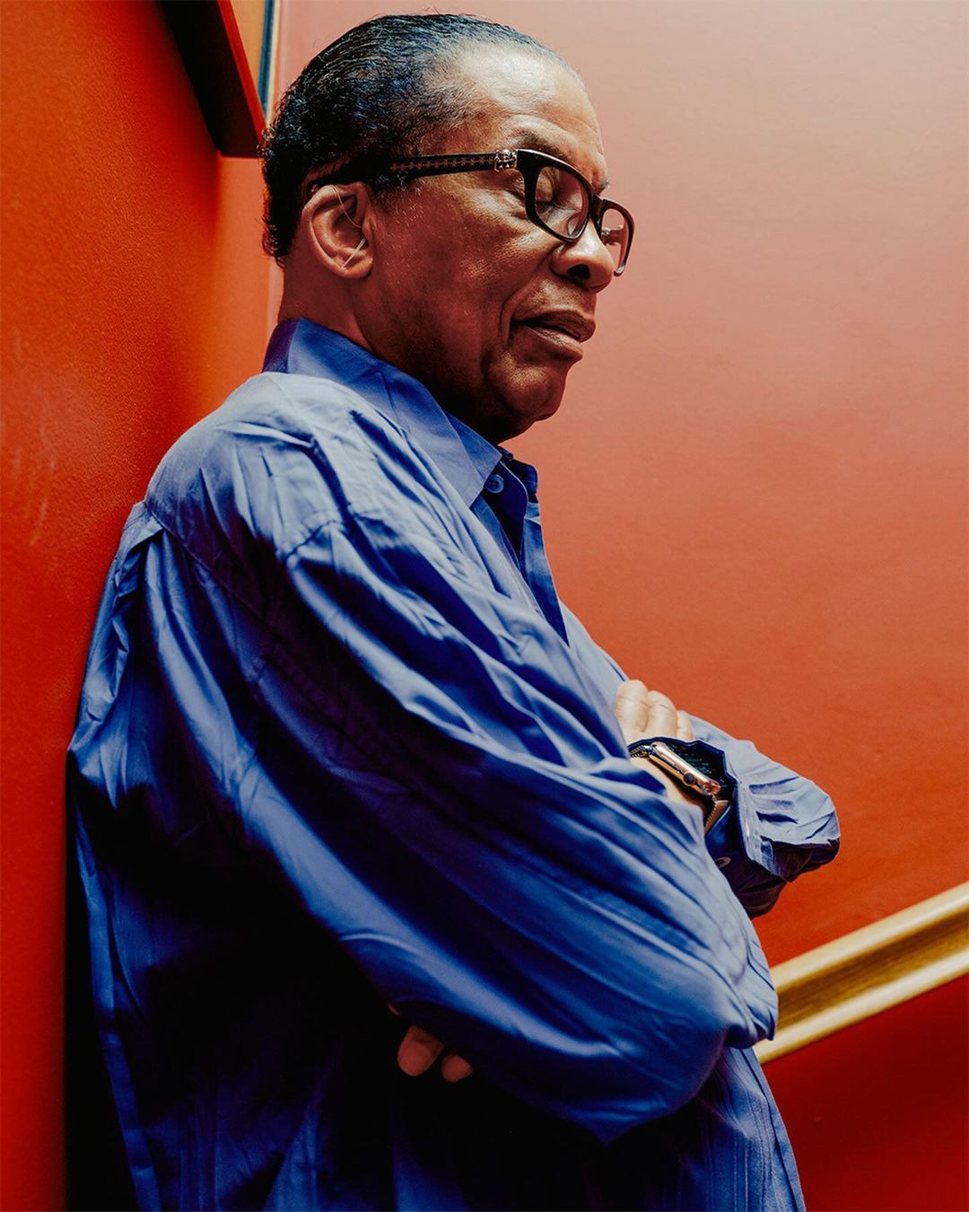 Portrait of an African-American man wearing glasses and a blue shirt with arms crossed, standing against a red wall.