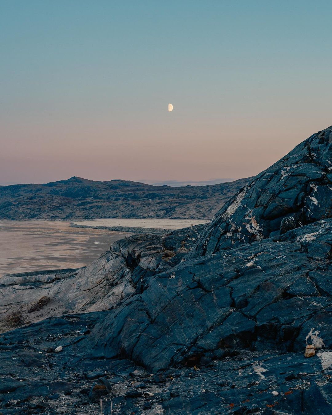 Dramatic landscape view featuring a rocky terrain foreground, calm water, distant hills, and a crescent moon against a blue sky at twilight.