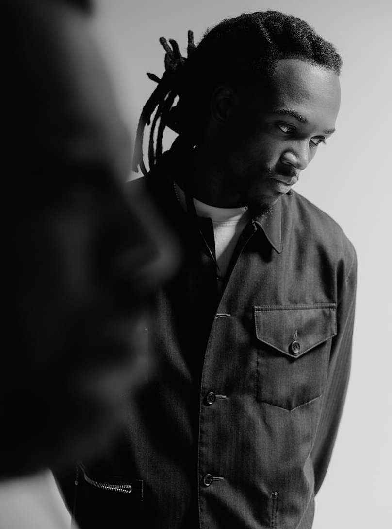 Monochrome portrait of a young African-American man with dreadlocks wearing a denim jacket, gazing off-camera with a blurred face in the foreground.