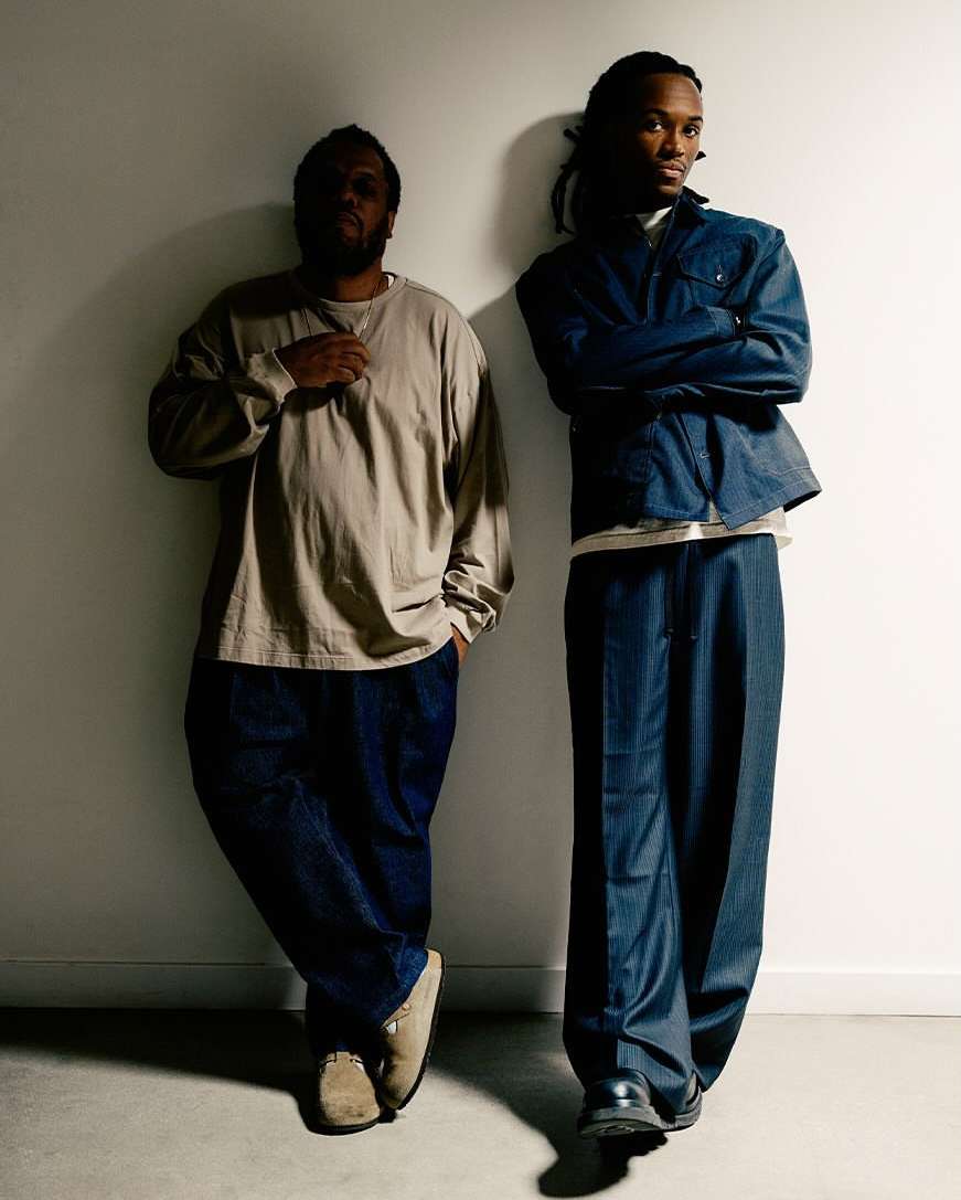 Two Black men pose against a white wall. One man wears a denim jacket and trousers; the other, a neutral top and dark pants.