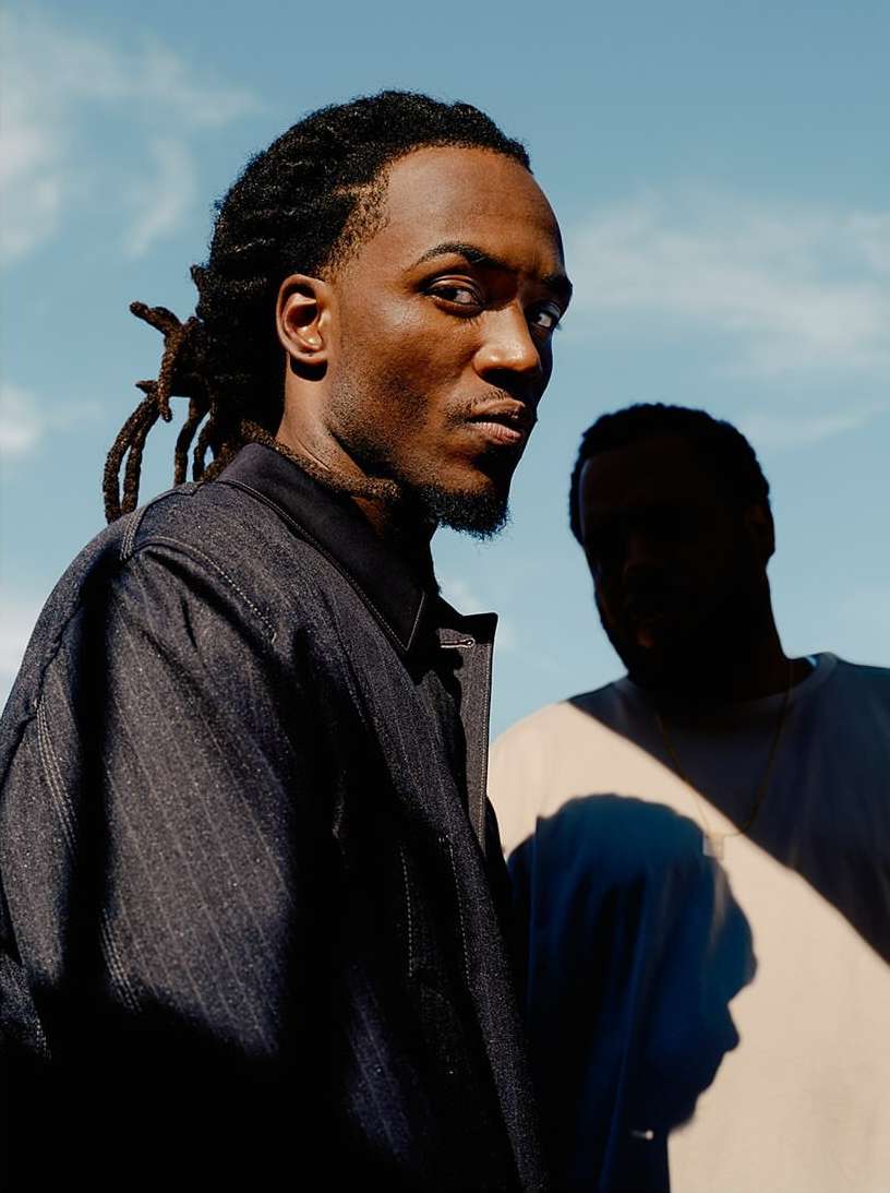Portrait of two African American men wearing jackets against a sky backdrop, captured with natural lighting and shallow depth of field.