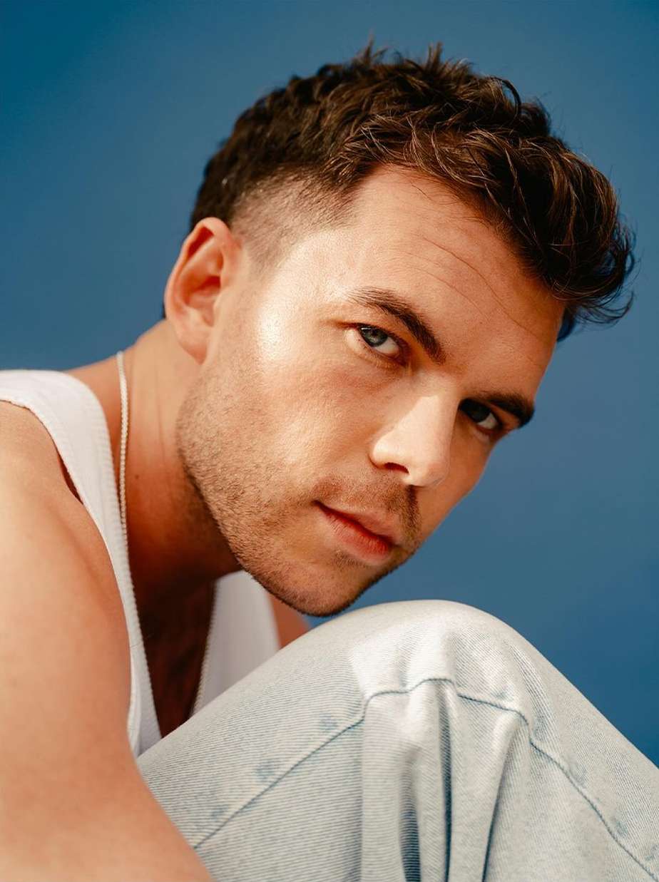 Portrait of a brunette male wearing a white tank top and light-wash jeans against a blue background, looking at the camera.