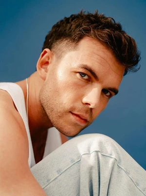Close up portrait of a man in a white tank top and light denim jeans against a solid blue background photographed by Daniel Dorsa