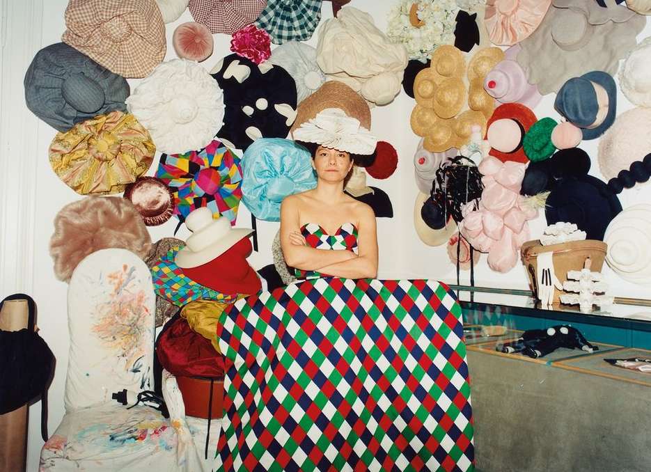 Portrait of an adult woman wearing a hat, standing in front of a wall of hats and a table covered with a patterned cloth.