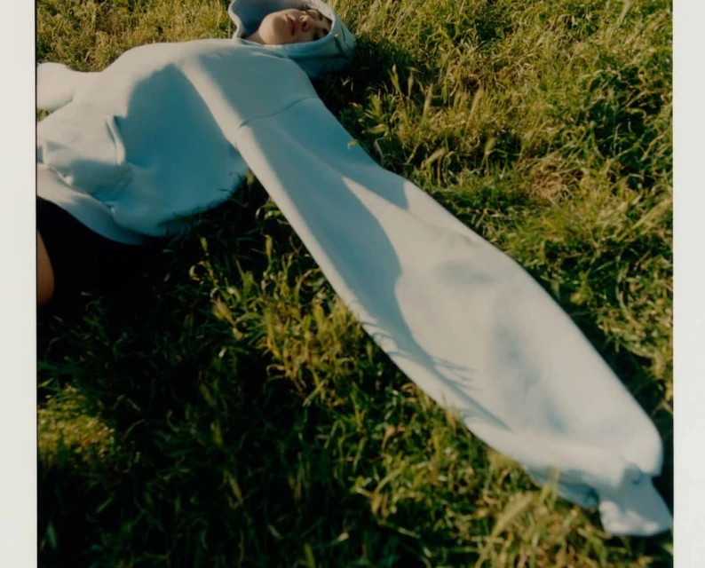 Woman in light blue hooded sweatshirt lies on her back in green grass in an outdoor setting on a sunny day, captured in an eye-level shot.