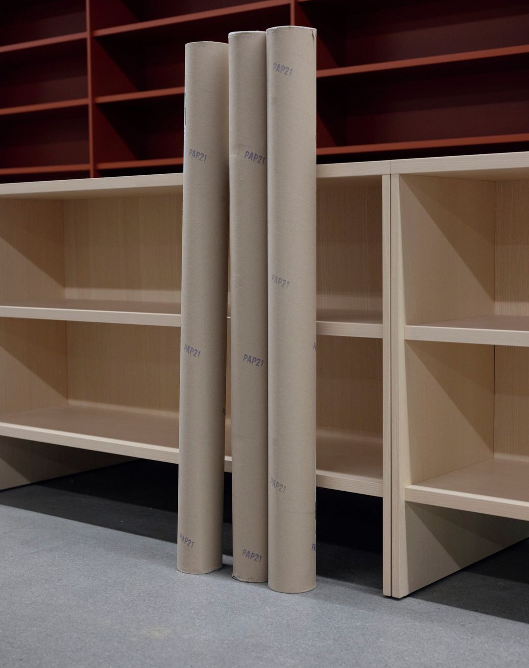 Three cardboard tubes stand upright against light-colored shelving, with a burgundy shelving unit in the background against a gray floor.