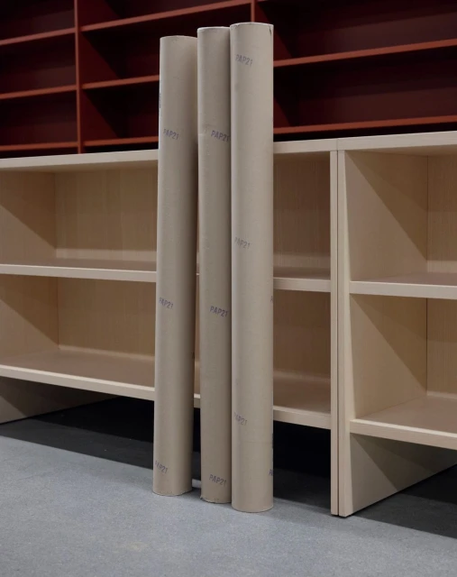 Three cardboard tubes stand upright against light-colored shelving, with a burgundy shelving unit in the background against a gray floor.