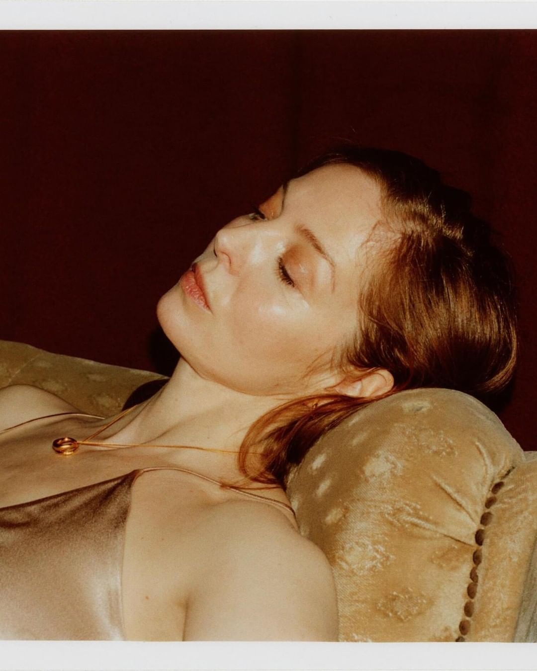 Woman lying on a plush sofa in soft lighting, showcasing a satin top and delicate gold necklace with a circular pendant.