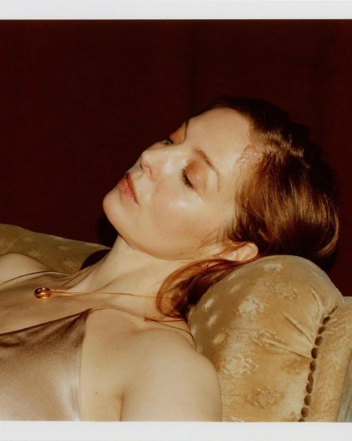 Woman lying on a plush sofa in soft lighting, showcasing a satin top and delicate gold necklace with a circular pendant.