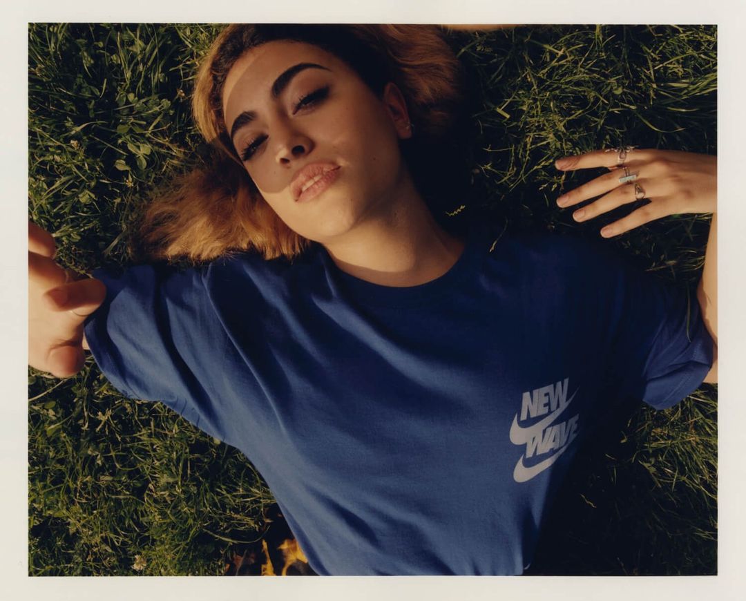 A young adult female with fair skin and light-brown hair lies on her back in green grass, wearing a blue t-shirt in a medium shot outdoors.