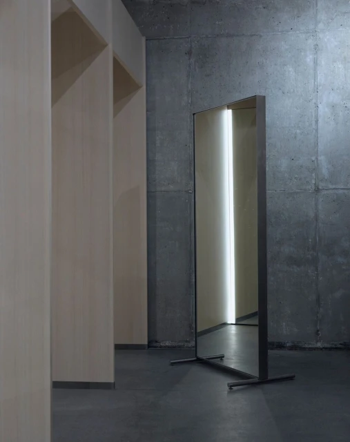 Modern interior featuring a full-length mirror with a metal frame against a textured concrete wall, reflecting a bright light source.