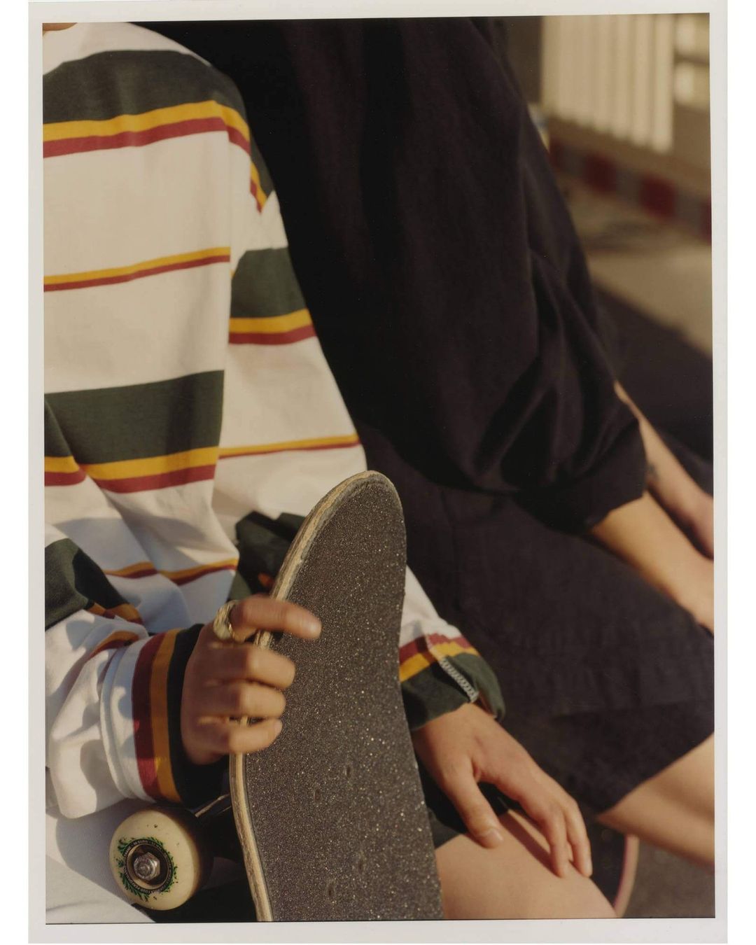 Two people sit on a ledge; one is holding a skateboard. The person in the foreground wears a striped shirt, with sunlight on their arm.