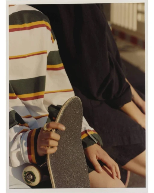 Two people sit on a ledge; one is holding a skateboard. The person in the foreground wears a striped shirt, with sunlight on their arm.