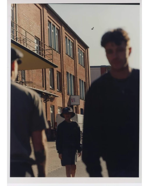 Three people walk on a street lined with red brick buildings on a sunny day, captured at eye-level in a medium shot.