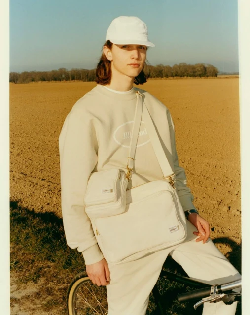 A young adult poses in a field wearing a white cap, light green sweatshirt and pants, and a white messenger bag with a bicycle nearby.