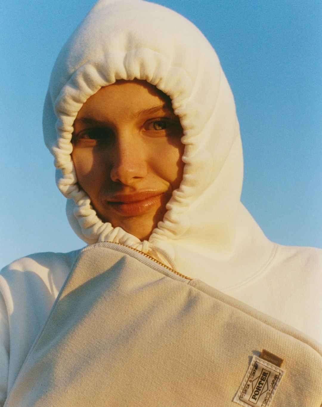 Woman in white hoodie holds tan laptop case against a bright blue sky in a close-up portrait, showcasing simplicity and modern design.