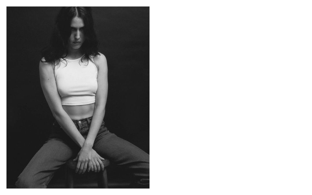 Black and white studio portrait of a young adult woman in a white crop top and jeans, seated on a stool against a dark backdrop.