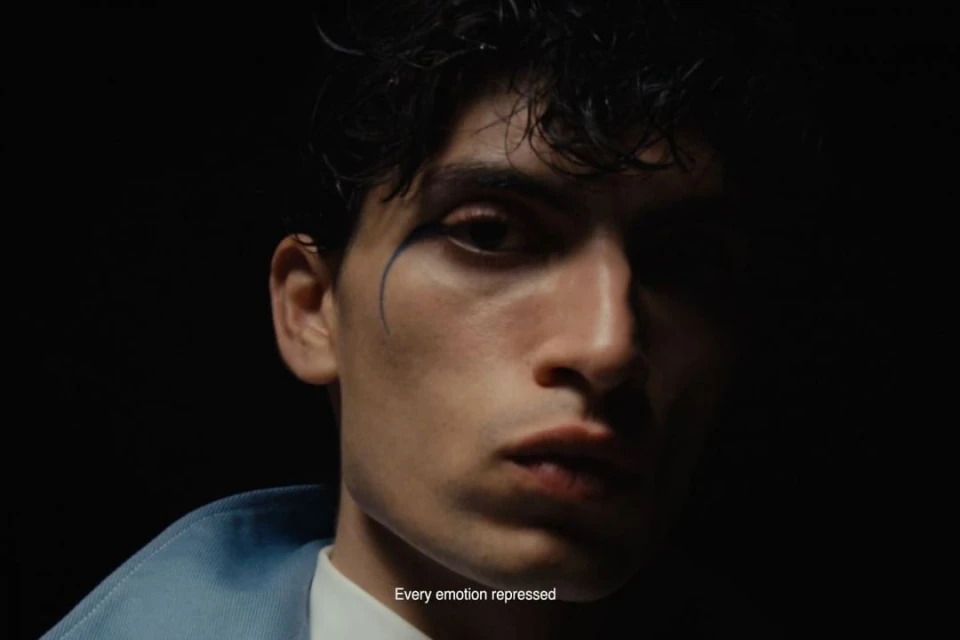 Close-up studio portrait of a young adult man with dark hair and blue eyeshadow against a black background wearing a light blue jacket.