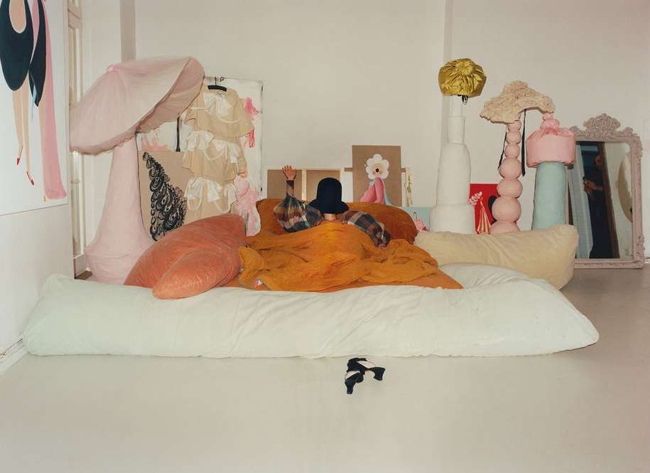 Whimsical fashion photo in a white room featuring an adult wearing a hat under an orange blanket, surrounded by pastel-colored props.