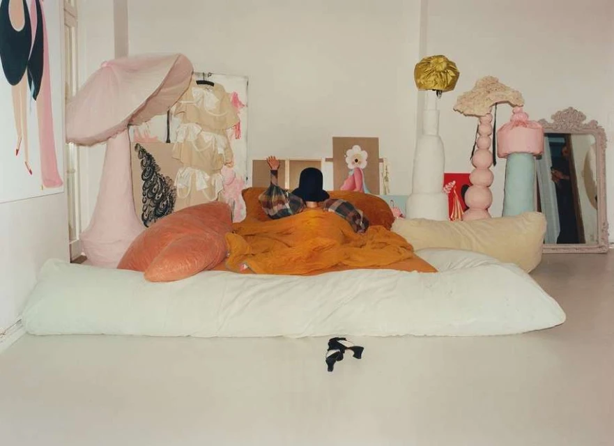 Whimsical fashion photo in a white room featuring an adult wearing a hat under an orange blanket, surrounded by pastel-colored props.