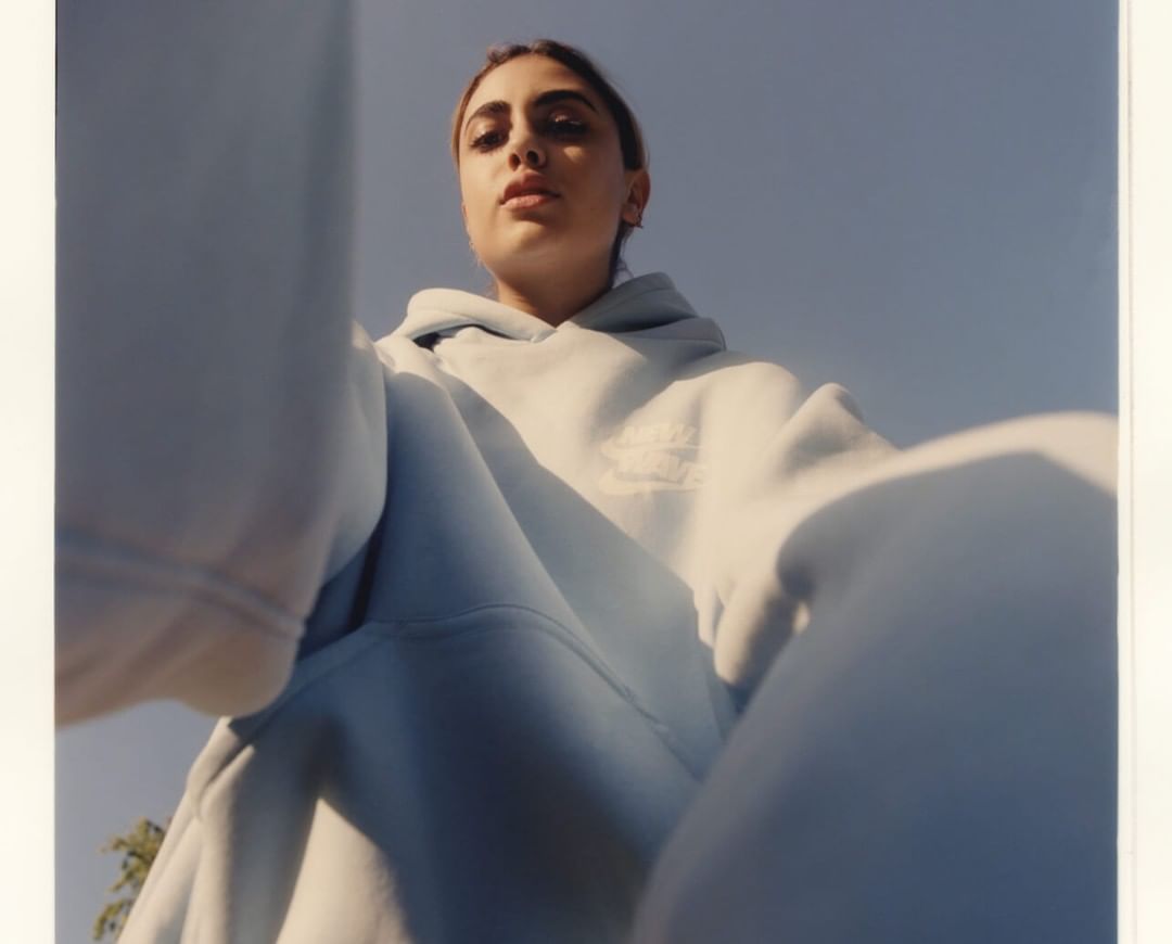 Low-angle shot of a young woman in a light blue hooded sweatshirt against a blue sky. The image has a vintage photograph aesthetic.