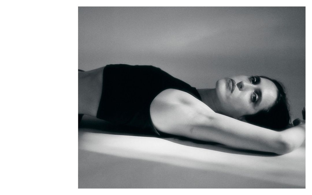 Monochrome portrait of a woman lying down with her arms above her head, soft lighting, and a contemplative facial expression.