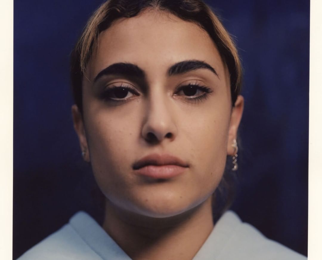 Close-up portrait of a woman with dark hair pulled back, light skin, and neutral expression against a dark blue background.
