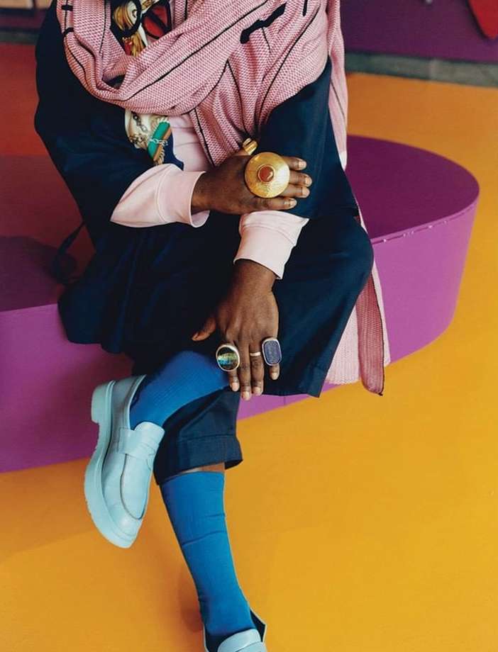 Stylish person sits on purple seating wearing rings, a scarf, and light blue shoes against a colorful background with blue socks.