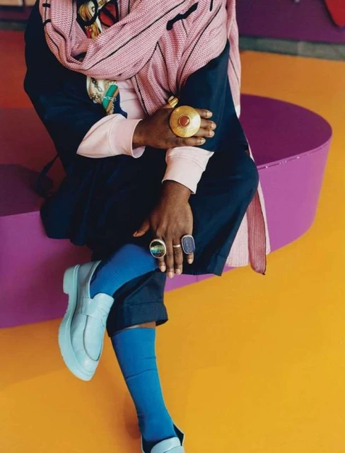 Stylish person sits on purple seating wearing rings, a scarf, and light blue shoes against a colorful background with blue socks.