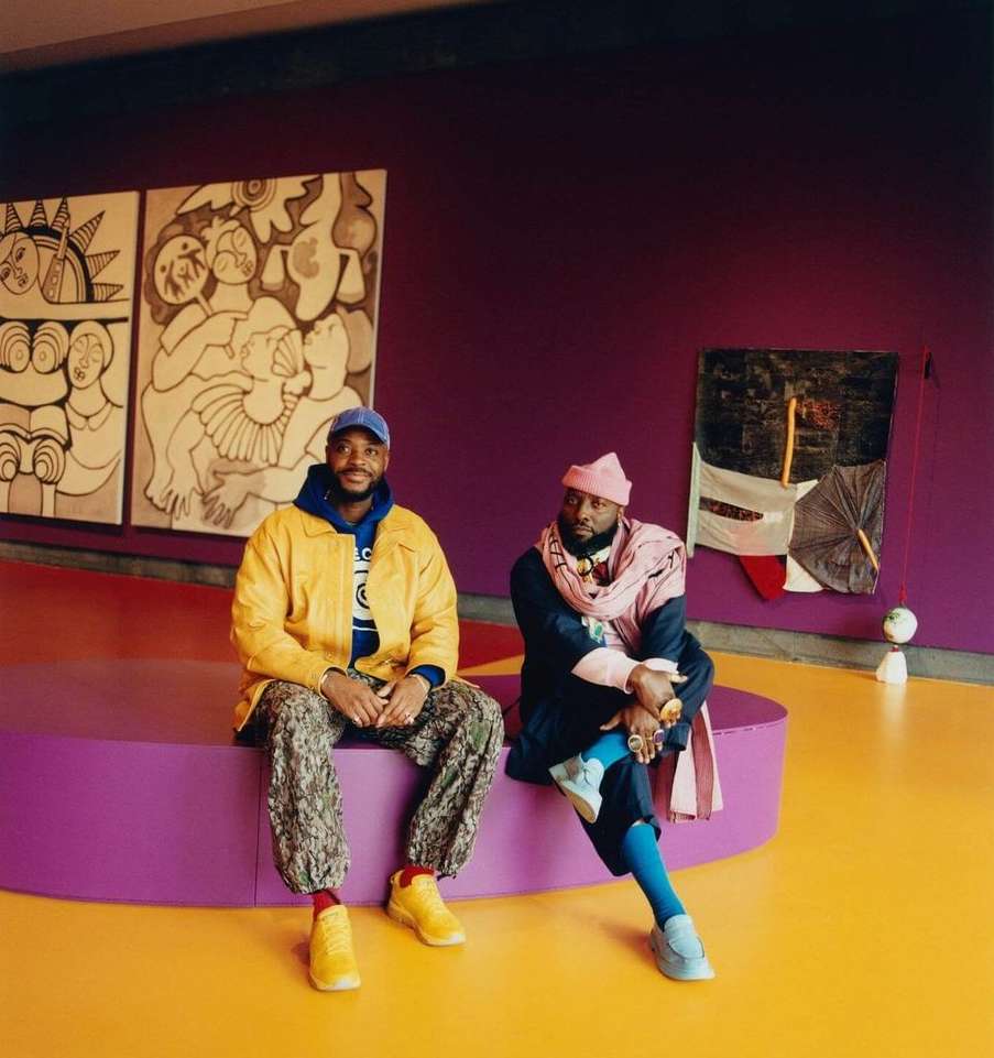 Two adult men pose seated on a purple bench in front of modern art; one wears yellow and tan, the other pink and navy.