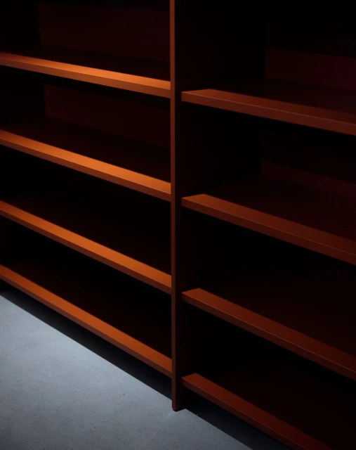 Empty shelves with red-brown wood finish in an indoor space; the shelves are evenly spaced on a light grey floor.