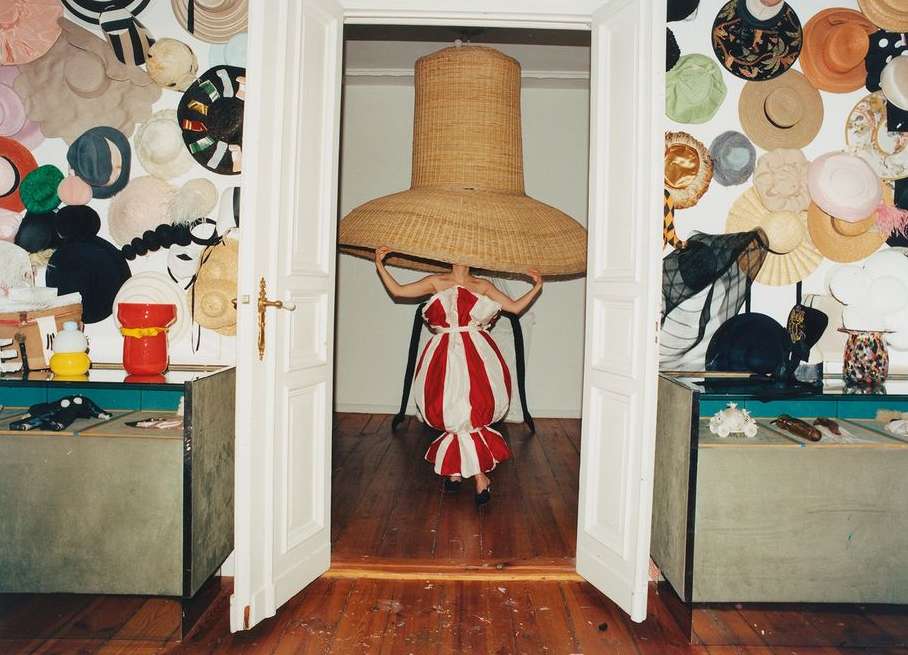 An adult female stands in a doorway wearing a white-and-red dress and a massive woven hat, with walls decorated with hats in the background.