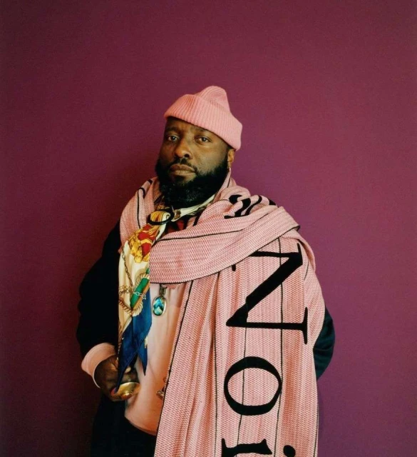 Adult male poses against a purple backdrop, wearing a pink beanie and scarf with black lettering, along with various jewelry.