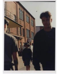 Three people walking down a city street alongside a multi-story red brick industrial building under a clear daytime sky.